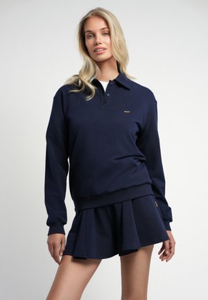 Vrouw draagt een donkerblauw poloshirt met lange mouwen en een bijpassende geplooide rok, staand tegen een effen lichte achtergrond.