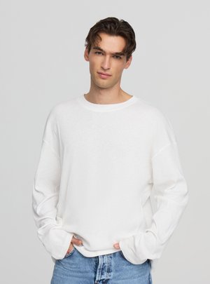 Giovane uomo con capelli castani che indossa una camicia bianca a maniche lunghe e jeans blu, in piedi contro uno sfondo chiaro e uniforme.