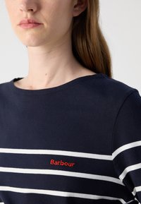 Tumesinane puuvillane topp, millel on horisontaalsed valged triibud ja väike punane "Barbour" logo rinnal. Ümar kaelus ja sile tekstuur.