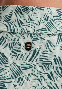 Camiseta polo con estampado de motivos de hojas verdes y azul verdoso sobre un fondo claro, con un logo negro de PME Legend adjunto.