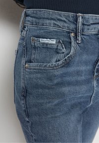 Modré džíny z denimu s mírným vyblednutím, designem s pěti kapsami a malou značkovou nálepkou na zadní kapse. Obsahují hardware ve zlatém tónu.