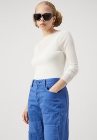 Bijela rebrasta bluza dugih rukava i plave cargo hlače s džepovima. Model nosi XXL crne sunčane naočale. Glatke teksture i jasne boje.