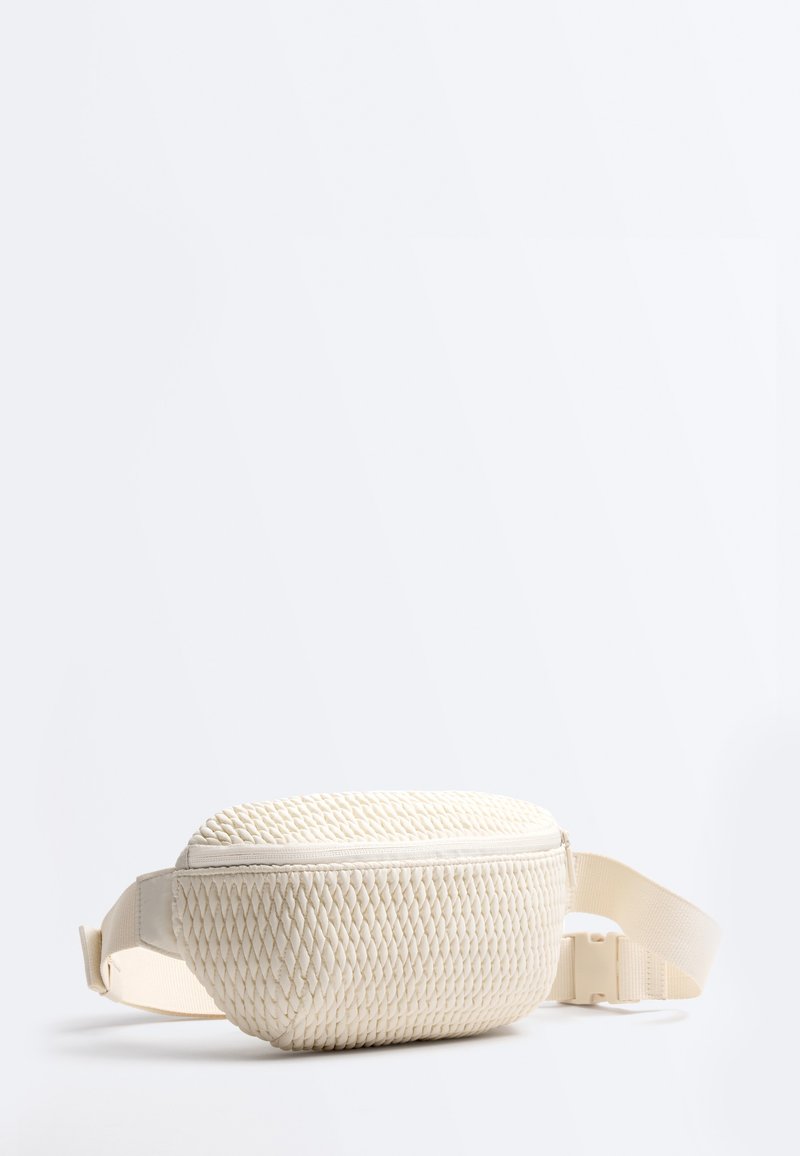 Petit sac banane beige avec face texturée, fermeture éclair et sangle ajustable avec boucle en plastique sur fond uni.