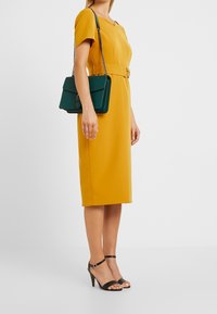 Gul knälång klänning med korta ärmar och figursydd midja, i kombination med en grön handväska med kedjeslinga och svarta klacksandaler.