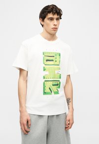 Νέος άνδρας με σκούρα σγουρά μαλλιά, φοράει λευκό T-shirt Nike Air Max με μεγάλο πράσινο γραφικό κείμενο και γκρι φόρμα.