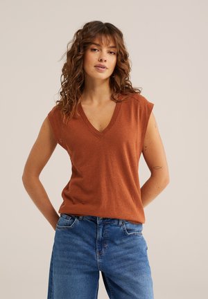 Donna con capelli castani ondulati che indossa un top senza maniche color ruggine con scollo a V e jeans blu, in piedi con le mani dietro la schiena.