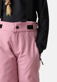 Różowe spodnie narciarskie z regulowanymi czarnymi szelkami, kieszeniami bocznymi i małym logo. Materiał wydaje się gładki i odporny na warunki atmosferyczne.