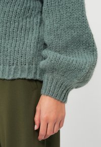 Primo piano di una mano appoggiata accanto a pantaloni verde oliva, che indossa una manica di un maglione lavorato a maglia leggero blu chiaro.