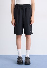 Person trägt schwarze Nike-Shorts, weiße Wadenlange Socken und schwarze Slip-On-Schuhe und steht auf einem blauen Boden vor einer weißen Wand.