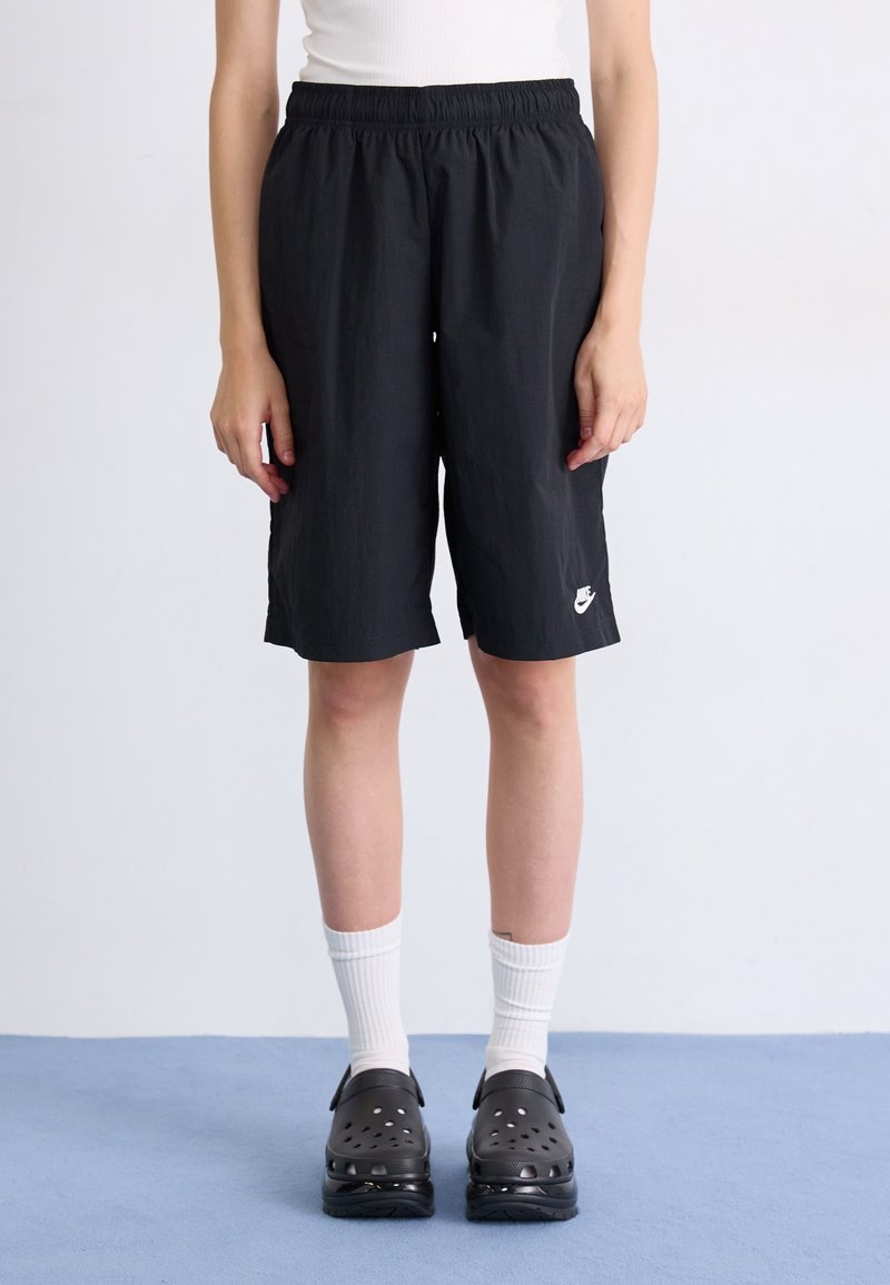 Person trägt schwarze Nike-Shorts, weiße Wadenlange Socken und schwarze Slip-On-Schuhe und steht auf einem blauen Boden vor einer weißen Wand.