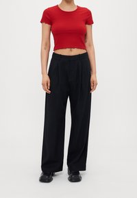 Kobieta w czerwonym, krótkim, prążkowanym topie, w luźnych, czarnych spodniach z wysokim stanem i czarnych, masywnych butach na jednolitym tle.