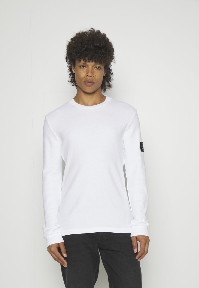 Personne aux cheveux bouclés portant une chemise blanche à manches longues avec une petite tache noire sur la manche gauche et un pantalon noir, debout devant un fond uni.