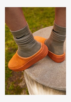 Des chaussures orange à enfiler en matériau texturé avec des détails perforés, associées à des chaussettes beiges à côtes avec un logo noir.