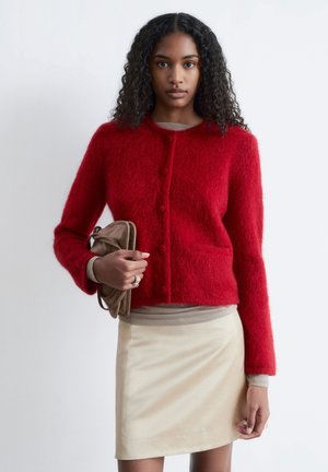 Roter, flauschiger Cardigan mit rundem Ausschnitt und fünf Knöpfen, getragen über einem beigefarbenen Oberteil. Das Modell hält eine beige Handtasche, kombiniert mit einem hellen Rock.