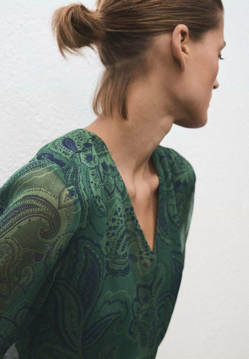 Blusa verde con estampado y escote en V, con mangas translúcidas, que presenta intrincados diseños de paisley en tonos de verde oscuro y azul.