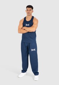 Mørkeblå atletisk tanktop med ribbet tekstur og hvidt logo, parret med matchende sweatpants med logo. Skoene er hvide.