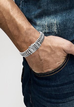 Silver chain bracelet with interlocking links, featuring a rectangular metallic clasp. Worn on a hand above dark denim jeans.