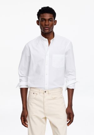 Homme portant une chemise blanche à boutons avec les manches retroussées et un pantalon beige clair, debout devant un fond blanc uni.