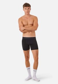 Shirtless man standing with arms crossed wearing black boxer briefs and white mid-calf socks against a plain white background.