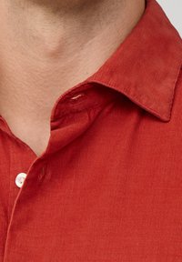 Camicia di velluto a coste rossa con colletto a bottone. Vista ravvicinata che mostra il tessuto testurizzato e il dettaglio del bottone bianco sulla pattina.