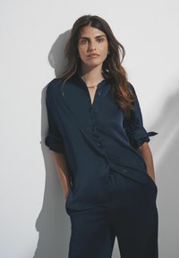 Navy blue button-up shirt, valmistettu sileästä kankaasta, jossa on käännetyt hihat ja rento istuvuus, yhdistettynä yhteensopiviin housuihin.