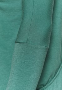 Teal långärmad tröja med en jämn, mjuk textur och något åtsittande ärmar, med en sömlös design vid axel och manschett.