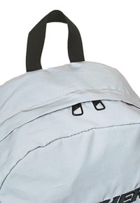 Mochila gris con un asa superior, cierres de cremallera y detalles negros. Fabricada con un tejido suave con costuras finas y una superficie texturizada.