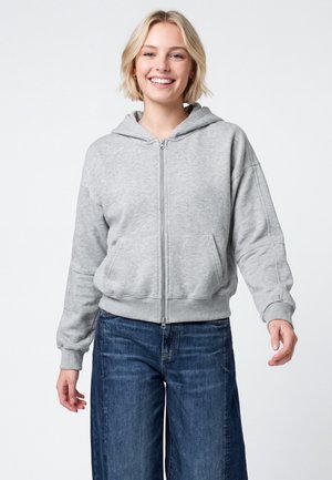 Ung kvinde med kort blondt hår smilende, iført en lysegrå hættetrøje med lynlås og viddeblå jeans, stående foran en hvid baggrund.