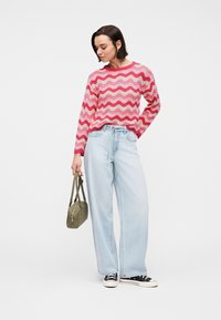 Γυναίκα που φοράει ροζ πουλόβερ με κυματιστό μοτίβο και ανοιχτόχρωμο μπλε τζιν παντελόνι με ευρύ πόδι, κρατώντας μια τσάντα ώμου σε ελαιόχρωμο χρώμα, στέκεται σε λευκό φόντο.