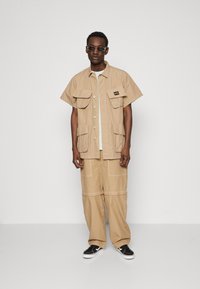 Tan button-up shirt with two front pockets, paired with wide-leg tan pants. Black sneakers and dark sunglasses complete the look.