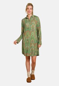Green paisley-patterned shirt dress with long sleeves, collar, and button placket; wool socks and brown slippers complete the look.