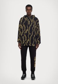 Must musta kulla kettide ja tekstimustriga, kombineeritud mustade dressipükstega, millel on kollane "COUTURE". Mustad sportijalatsid lõpetavad ilme.
