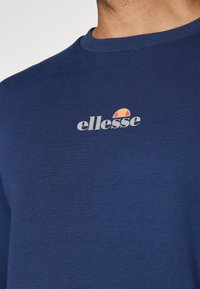 Granatowa bawełniana koszulka z okrągłym dekoltem, z białym logo "ellesse" oraz małym, multikolorowym emblematem nad nim.