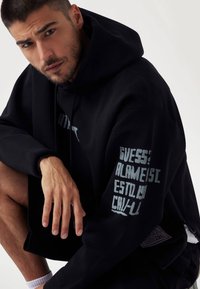 Černá mikina s kapucí v oversized střihu, zdobená modrým grafickým textem na rukávech a detaily se zipem. Hladká textilie.