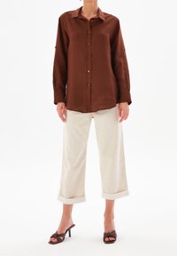 Brown linen button-up shirt with a collar, long sleeves, and wooden buttons; paired with light beige cropped pants and dark brown heeled sandals.