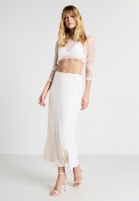 Una donna con un crop top in pizzo bianco, una gonna midi in satin a vita alta e sandali bianchi con tacco alto, in piedi davanti a uno sfondo neutro.