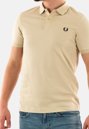 Polo beige avec un tissu texturé, à manches courtes, patte de boutonnage à deux boutons, et un logo brodé noir sur le côté gauche de la poitrine.