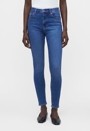 Femme portant un jean skinny bleu taille haute associé à des ballerines noires et un top blanc sans manches, debout devant un fond uni.