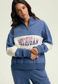 Niebieska bluza z półzamkiem, z białym pasem i różowym napisem "MICHIGAN". Prążkowane mankiety i dół nadają fakturę.