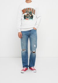 Man i vit sweatshirt med en fisk och texten "Montana Big Country", trasiga blåjeans och röda sneakers, stående mot en vit bakgrund.