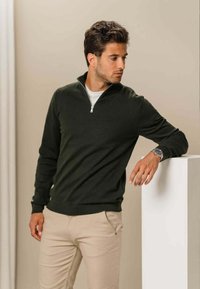 Mørkegrøn pullover med kvart lynlås i halsen, ribbede manchetter og kant. Båret med beige bukser, sat mod en neutral baggrund.