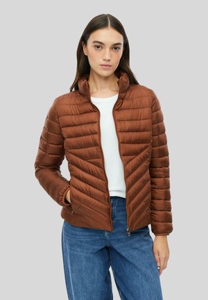 Frau trägt eine braune gesteppte Jacke mit Reißverschluss über einem weißen Oberteil und blauen Jeans vor einem grauen Hintergrund.