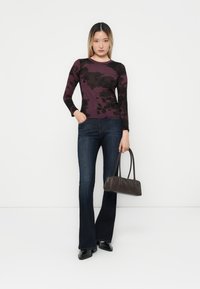 Langsleeve top in donkerpaars met zwart abstract patroon, gecombineerd met donkerblauwe flared jeans en een kleine donkergrijze handtas.