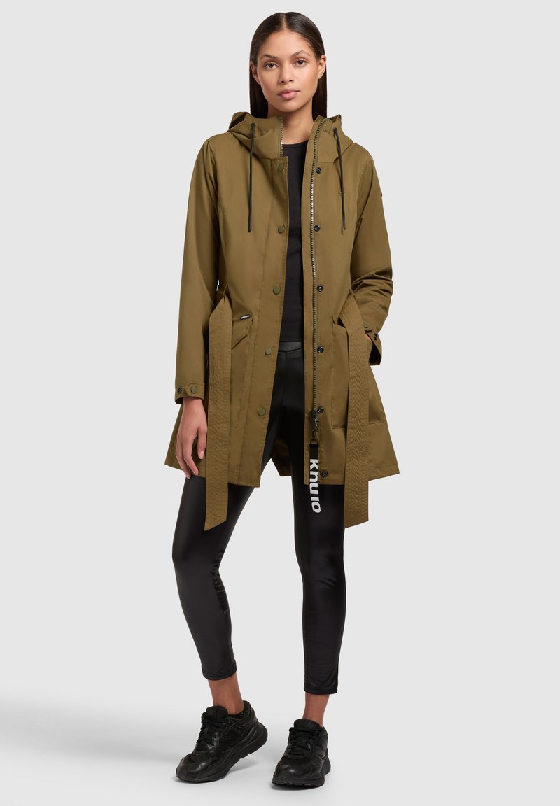 Olivgrüne Kapuzenjacke mit vorderem Knopfverschluss, seitlichen Taschen, verstellbaren Kordeln und einem lockeren Schnitt, kombiniert mit schwarzen Leggings und Sneakers.