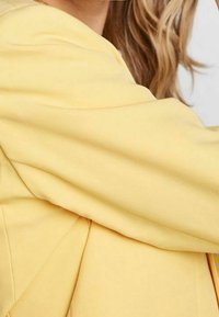 Żółty materiał o gładkiej fakturze, charakteryzujący się luźnym krojem i zdobieniami. Zbliżenie na rękaw i szczegóły po bokach.