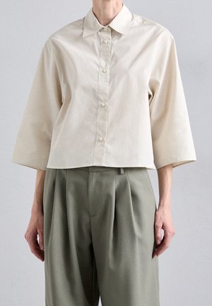 Person iført en løs cremefarvet knap-op skjorte med trekvartærmer og højtaljede vidde bukser i olivengrøn.