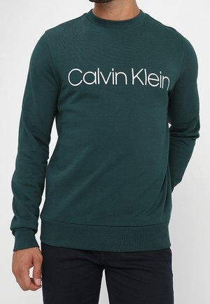 Homme portant un sweat-shirt à manches longues vert foncé avec "Calvin Klein" imprimé en blanc sur la poitrine, associé à un pantalon noir.
