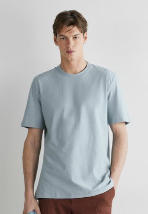 Jonge man met kort bruin haar, gekleed in een lichtblauw T-shirt met korte mouwen en roestkleurige broek, staand tegen een witte muur.