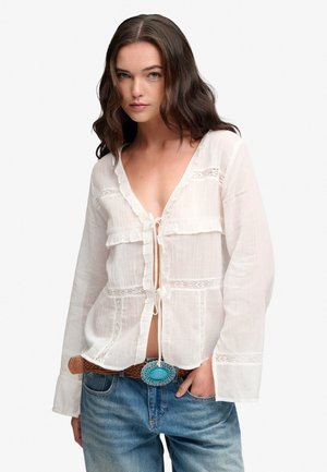 Mujer con cabello castaño ondulado que lleva una blusa blanca transparente con lazos y vaqueros azules con un cinturón marrón con una gran hebilla turquesa.