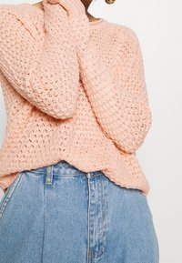 Vaaleanpunainen neulepusero, jossa on väljää ja ilmavaa rakennetta, pitkät hihat ja pyöreä kaula-aukko, yhdistettynä korkeavyötäröisiin farkkushortseihin.